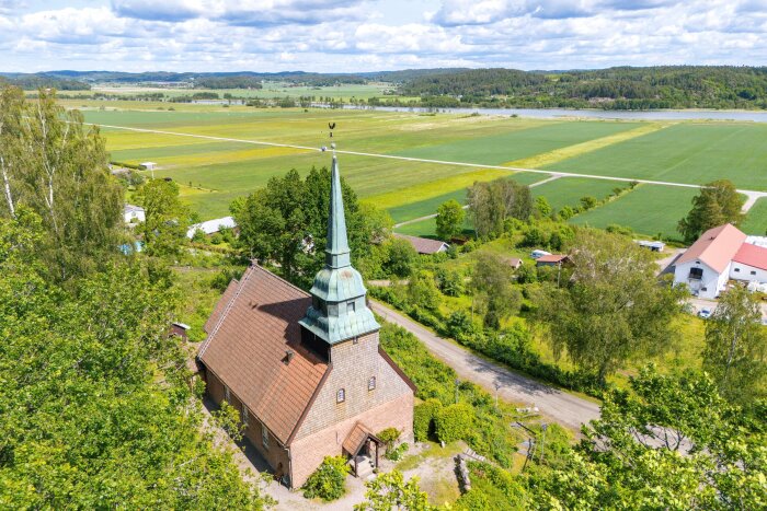 Flygfoto av en bostad med torn och klockspel omgiven av grönska och åkrar i ett lantligt landskap.