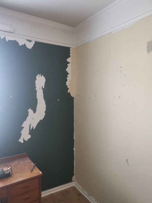 Vägg med bortsliten tapet i varierande skick, visar delvis exponerad mörk vägg. Träbord med verktyg synligt i hörnet.