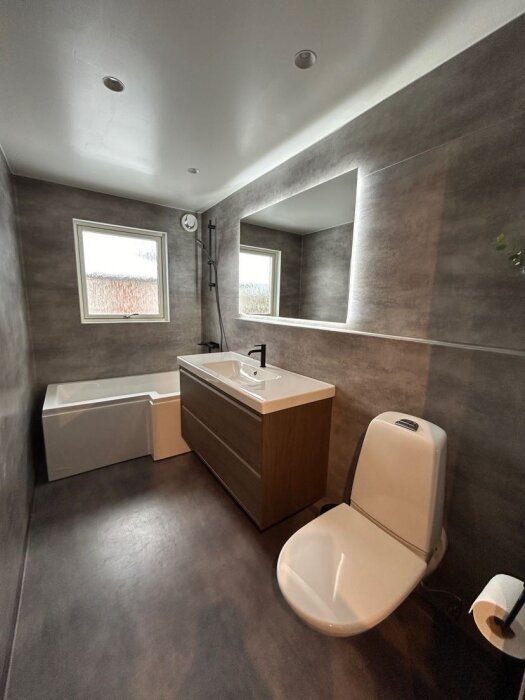 Modernt badrum med grå väggar, inbyggd badkar, vit toalett, handfat med trädetaljer och stor spegel ovanför.