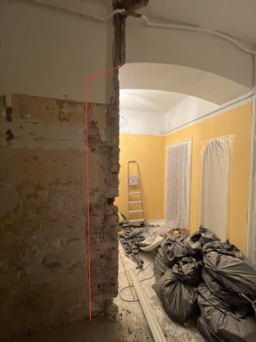 Tegelvägg där en del är markerad för borttagning, omgiven av byggmaterial och verktyg i ett rum med orange väggar och täckplast.