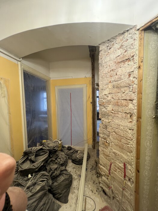 Renoveringsområde med exponerat tegelvalv och byggmaterial, inklusive svarta säckar och verktyg, visar borttagen sektion av en bärande vägg.