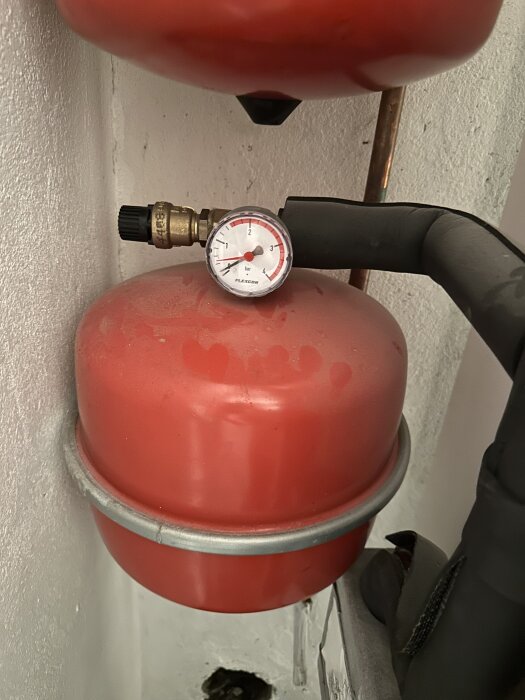 Röd trycktank med tryckmätare på 1 bar, ansluten till ett rör i en hemmiljö.