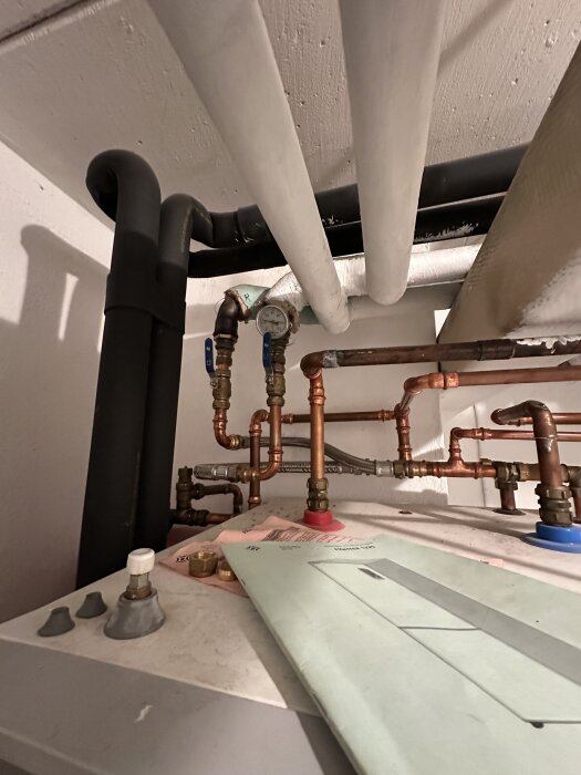 Rör och ventiler i ett tekniskt rum med kopparledningar och manometer. Ett dokument ligger på en maskin.