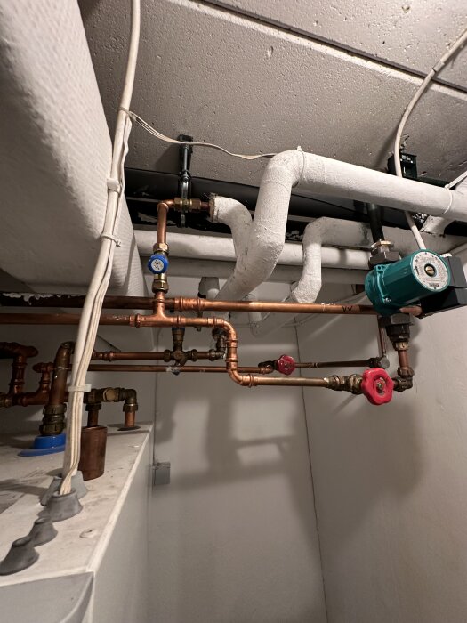 Kopparrör och ventiler i en källare, vit isolering runt rören och en grön pump synlig.