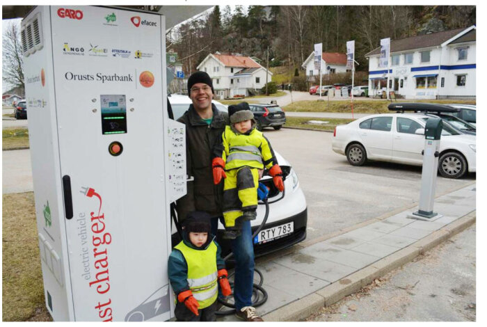 Man med två barn bredvid en laddstation för elfordon, en bil laddas i bakgrunden i ett bostadsområde.