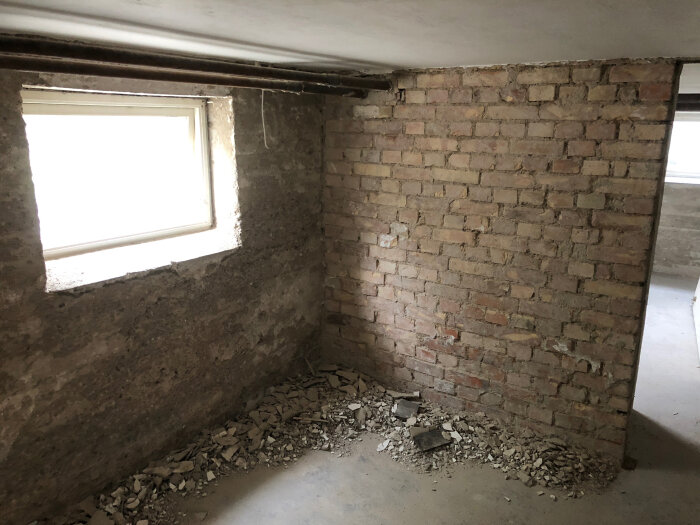 Källarrum med tegelvägg, trasigt ytskikt och skräp på golvet, intill ett litet fönster.