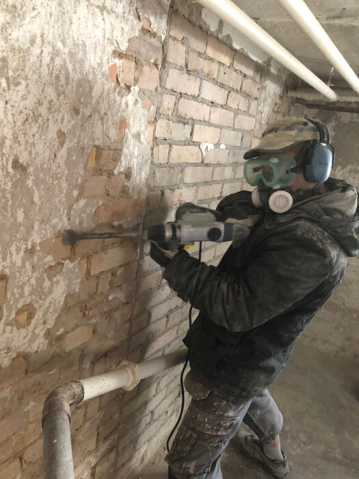 Person i skyddsutrustning använder bilmaskin på gammal putsad vägg i källare med synliga tegelstenar och rör.