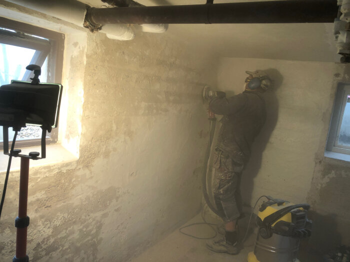 Person slipar en källarvägg med betongslip, maskiner och dammsugare syns.