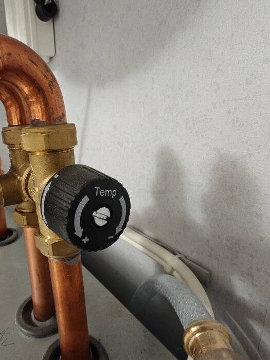 Vred för temperaturjustering på rör, märkt med "-" och "+", sitter på kopparrör i ett rum med vit vägg i bakgrunden.