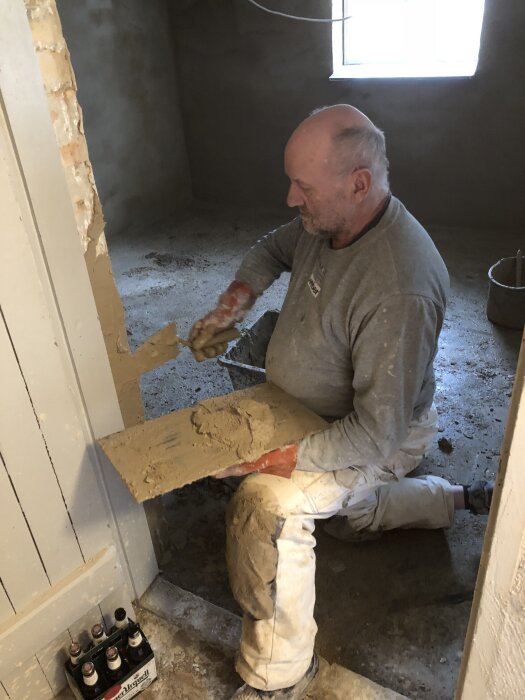 Murare putsar en vägg i en renovering av en källare, med verktyg och material synliga. Ljuset strömmar in från ett fönster i bakgrunden.