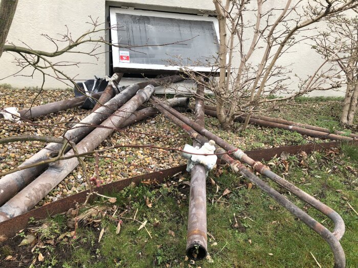 Gamla metallrör och grus framför en källarfönsteröppning, omgivna av gräs och buskar.