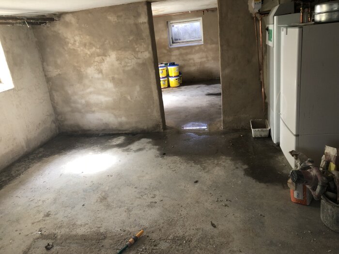 Renoverad källare med vita putsade väggar och betonggolv, vit panncentral synlig till höger och flera hinkar i bakgrunden nära ett litet fönster.