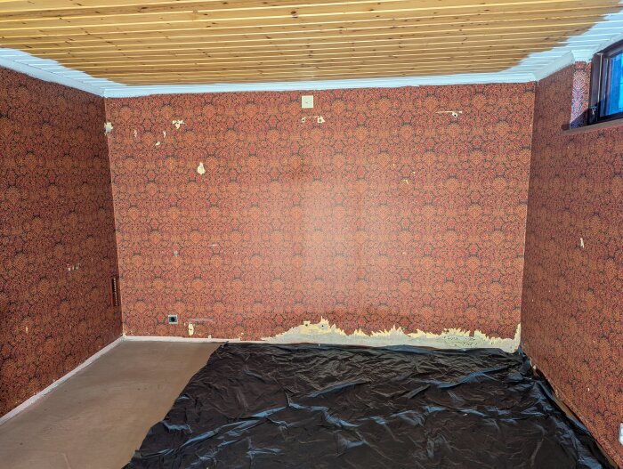 Rum med röd tapet, synliga fuktfläckar längst ner på väggen och plastskydd på golvet, tak i ljust trä.