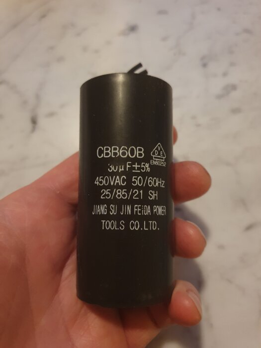 Svart kondensator märkt "CBB60B" och "30µF ±5%", specificerad för 450VAC och fabrikerad av "JIANG SU JIN FEI DA POWER TOOLS CO. LTD", hålls i en hand.