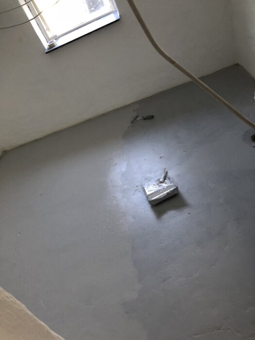 Nymålat golv i källare med grå färg, en roller ligger mitt på golvet. Ett fönster syns i bakgrunden, rummet verkar nymålat och renoverat.