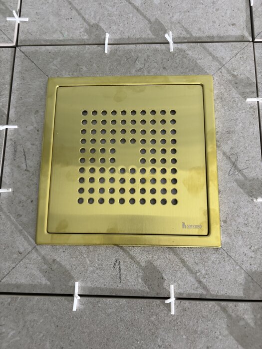 Guldfärgad golvbrunn monterad bland grå plattor vid kuvertskärning, med vita distanser för plattsättning.