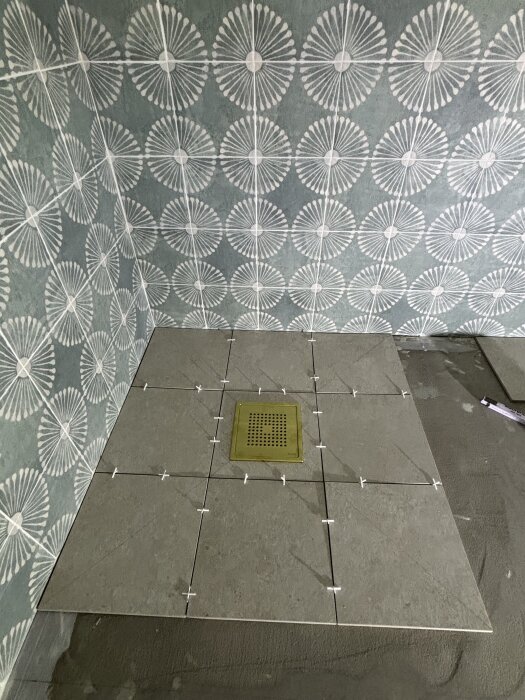 Plattsättning med kuvertskärning påbörjad i duschhörna med avloppsbrunn, turkose mönstrade väggar och oklara golvplattor, plattdelare synliga.