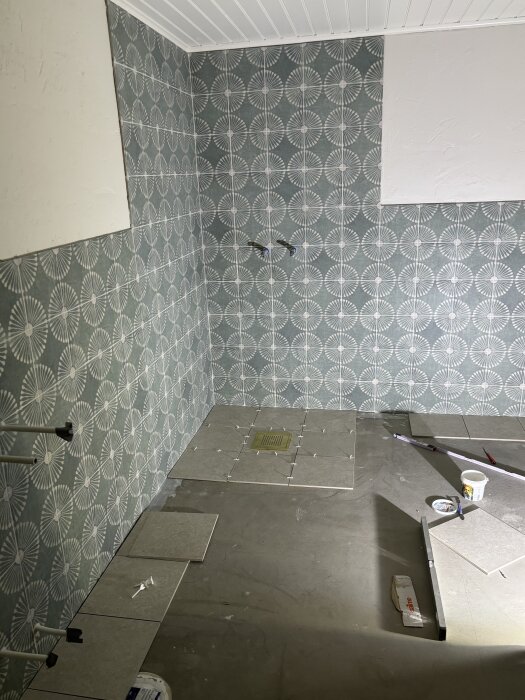 Vägg och golv i badrum under kakelinstallation med grå plattor och mönstrad vägg. Verktyg och material synliga.
