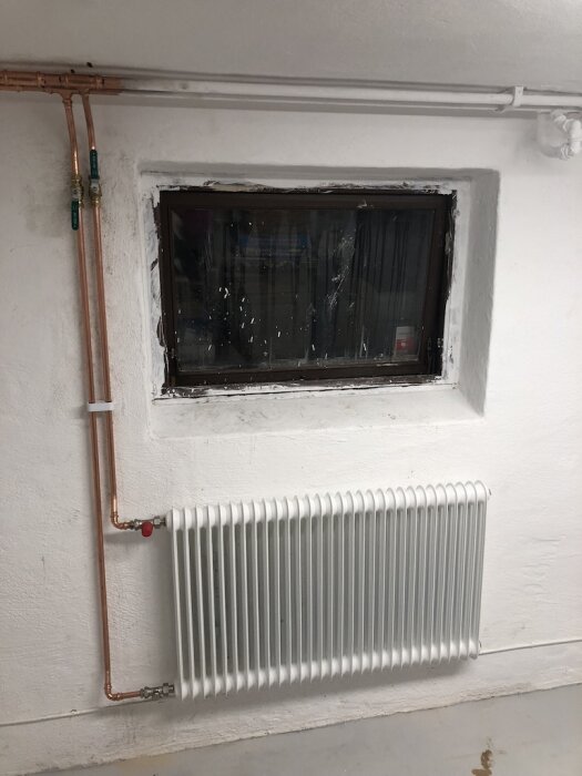 Vitt radiator installerad under ett källarfönster med kopparrör längs väggen, del av renoveringsprojekt i en tvättstuga.