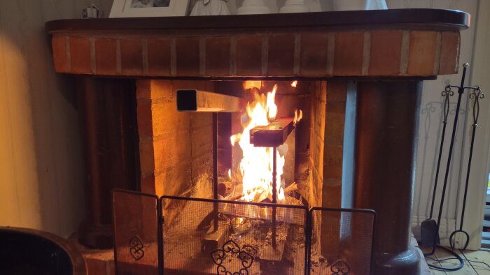 Brasa i en murad öppen spis med lågor som lyser upp eldstaden. Ved brinner inne i spisen med ett skyddande galler framför.