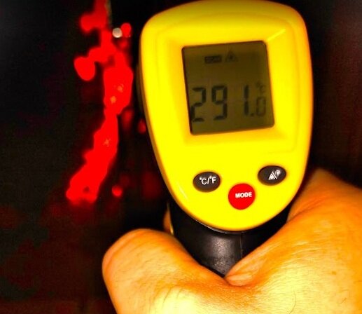 En hand håller en gul infraröd termometer som visar temperaturen 291 grader Celsius. Bakgrunden är oskarp med röda och svarta nyanser.