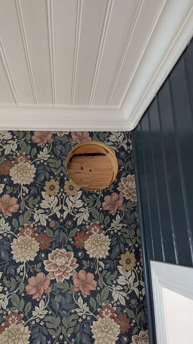 Hål i tapetserad vägg för imkanalinstallation, omgiven av blommigt mönster och vit takpanel.