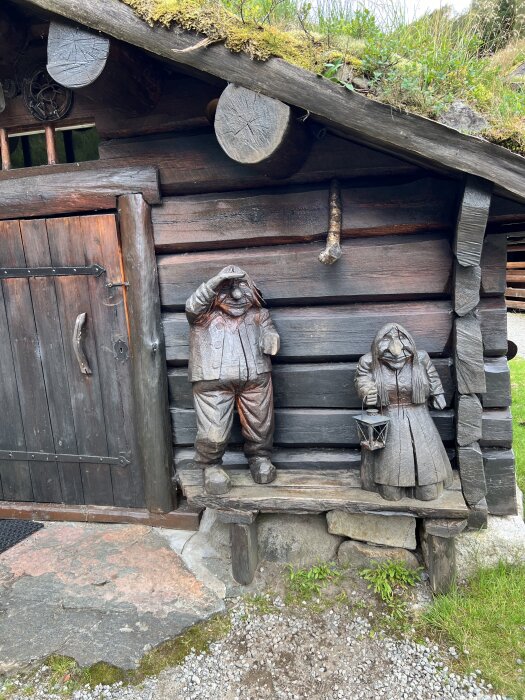 Träskulpturer av en man och kvinna framför en timrad stuga med mossbeklätt tak. Kvinnan håller en lykta, och mannen lyfter handen till pannan.
