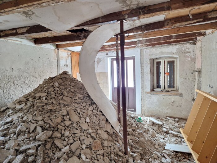 Rivningsarbete med hög av byggrester, trasigt tak och synliga reglar i ett rum med fönster och dörr i bakgrunden.