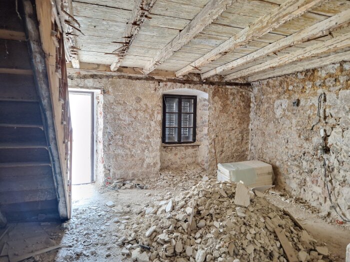 Rivet rum med synliga träbjälkar, stenväggar och en hög med bråte på golvet, fönster och dörröppning ut mot dagsljus.