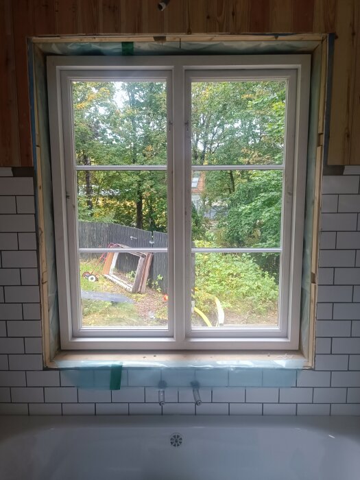 Fönster ovanför badkar med vita kakelplattor runtomkring, trä och tätning runt fönstret, utsikt mot träd och trädgård.