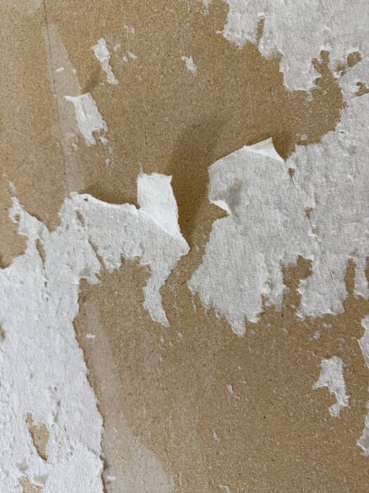 Vägg med pappersrester och skador efter borttagen vävtapet, synliga ojämnheter och fläckar på ytan.