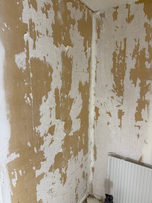 Vägg i förrådsrum med bortskrapad vävtapet, vita och bruna bitar av papprester kvar, synligt element nedanför.