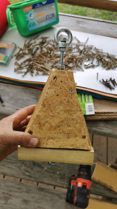 Fyrkantigt trätillbehör med ögla upptill, hålls av en hand. Bakgrund med spik och verktyg.