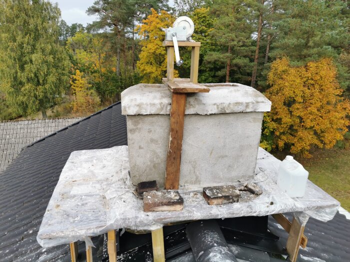 Chimney on a sloped roof with a makeshift wooden and plastic covering, surrounded by colorful autumn trees.