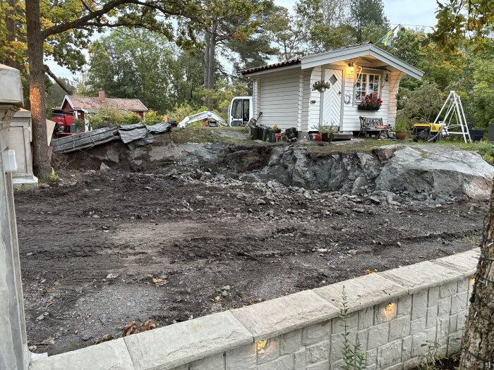 Uppfart med sprängd mark intill ett litet trähus i en trädgårdsmiljö. Bakom huset syns en röd byggnad och skog i bakgrunden.