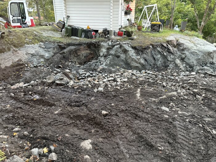 Uppfart efter sprängning med jord och mackadam, synliga berg och en grävmaskin vid ett hus med vit fasad och träd i bakgrunden.