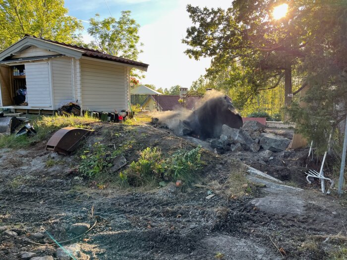 Ett litet uthus vid en uppfart, en hög med sprängda stenar, damm som stiger, och träd i bakgrunden i solnedgången.