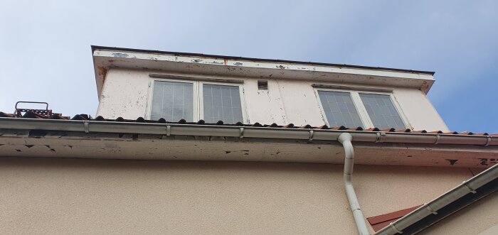 Takkupa med två fönster, slitna träpaneler och takränna, förbereds för renovering med ny papp och plåt.