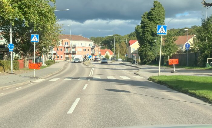 Trafikskyltar och omledningsskyltar vid en väg i ett bostadsområde med bilar och övergångsställe, grönt gräs och träd längs vägen.