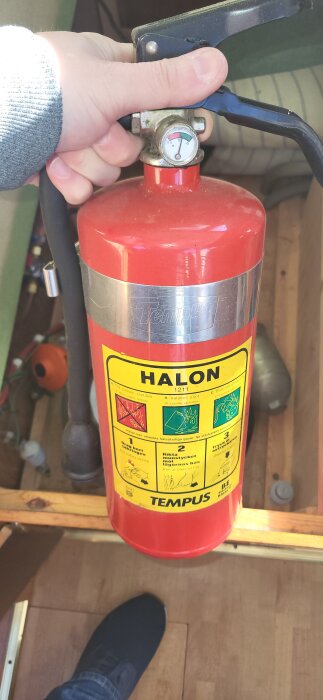 Röd brandsläckare av typen Halon hålls i en hand inomhus, märke Tempus syns tydligt på etiketten.