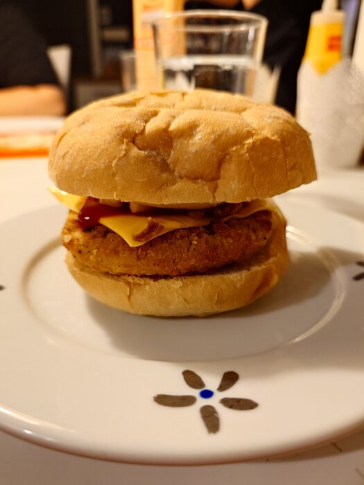 Crispy chicken-burgare med ost, lök, senap, ketchup och sourcreme pepper på en tallrik. Bakgrunden visar suddiga flaskor och ett glas vatten.
