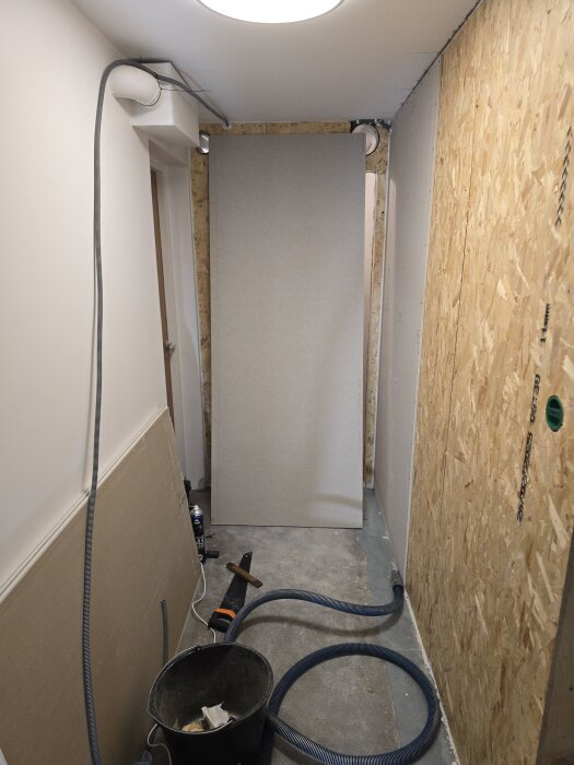 Korridor under renovering med OSB-skivor och gipsskiva på väggen, ventilationsrör i taket och verktyg samt dammsugarslang på golvet.