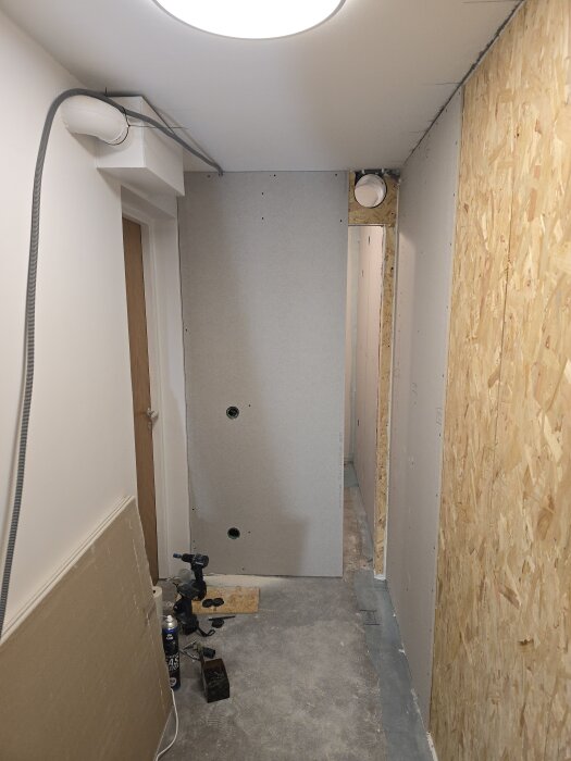 Korridor med gipsväggar, OSB-vägg, synligt ventilationsrör i taket och verktyg på golvet. Arbete pågår med att installera gipsskivor.