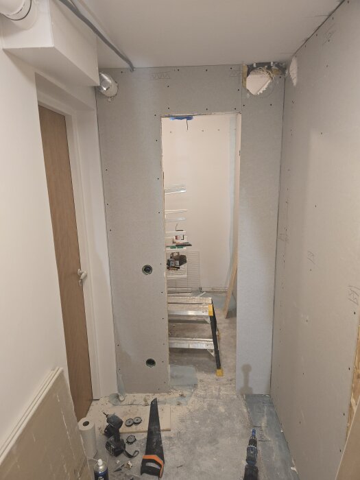 Renovering pågår med gips och två ventilationshål i en smal passage, verktyg på golvet och en stege i bakgrunden.