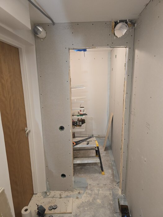 En halvfärdig vägg med gipsplattor monterade i en hall, synliga ventilationsrör och byggmaterial spridda på golvet.