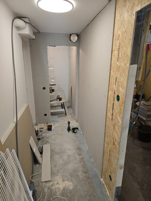 Renovering av källare, ny vägg med OSB och gips, synliga verktyg och ventilationsrör, korridor med ofärdigt golv och gipsmaterial på golvet.