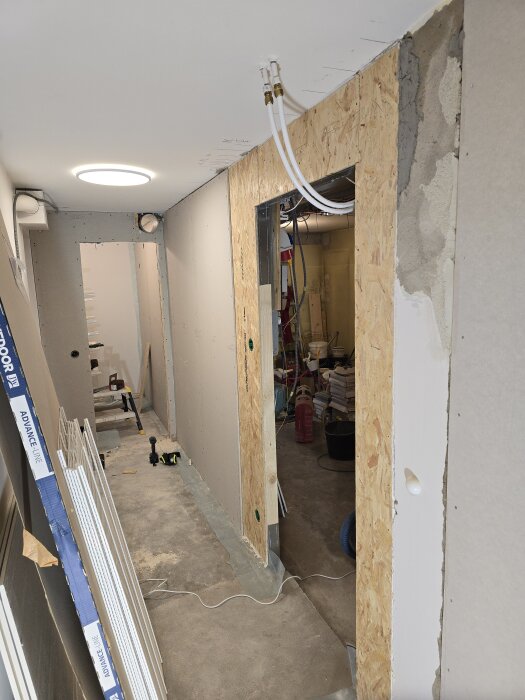 En korridor under renovering med reglad vägg, OSB-skivor och gipsskivor. Verktyg och material ligger på golvet. Synliga ventilationsrör i taket.