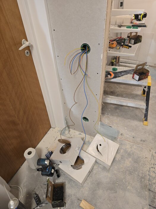 Gipsvägg med hål där elkablar hänger ut, omgivna av byggverktyg och material, bredvid en trädörr och hylla med verktyg i bakgrunden.