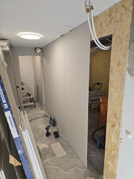Vägg under konstruktion med OSB- och gipsskivor. Elektriska verktyg på golvet och ledningar hängande från taket. Puts synlig vid dörrkarmen.