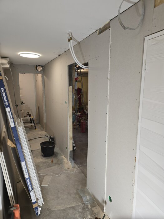 Korridor under renovering med gipsskivor på väggarna och synliga ventilationsrör i taket. Verktyg och material längs väggarna.